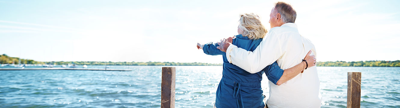 Older man and woman with arm around each other facing a lake, woman pointing outwards